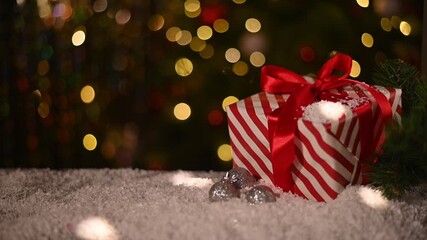 red and white striped Christmas gift with red ribbon sits next to silver disco ball ornaments on snowy surface. background soft bokeh lights from Christmas tree, adding festive touch. - Powered by Adobe
