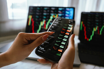 A person holding a smartphone showing stock market data with trading charts displayed on laptop screens in the background.
