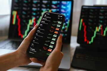 A person holding a smartphone showing stock market data with trading charts displayed on laptop screens in the background.
