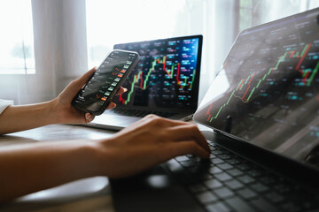 A person holding a smartphone showing stock market data with trading charts displayed on laptop screens in the background.