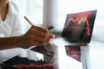A person holding a smartphone showing stock market data with trading charts displayed on laptop screens in the background.