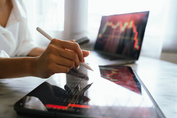 A person holding a smartphone showing stock market data with trading charts displayed on laptop screens in the background.