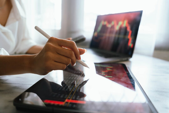 A person holding a smartphone showing stock market data with trading charts displayed on laptop screens in the background.