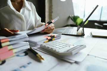 Close-up of businesswoman working at her desk with laptop and stack of documents, focusing on paperwork, reports and financial analysis in a busy office environment.