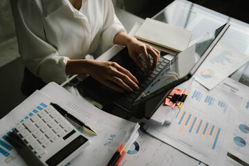 A person working with a calculator and financial documents on a desk, analyzing data from charts displayed on a laptop in a bright office environment.
