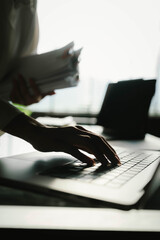 Close-up of businesswoman working at her desk with laptop and stack of documents, focusing on paperwork, reports and financial analysis in a busy office environment.