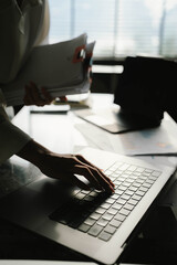 Close-up of businesswoman working at her desk with laptop and stack of documents, focusing on paperwork, reports and financial analysis in a busy office environment.
