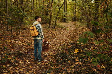 Man standing on forest trail with Australian Shepherd dog, beside them surrounded by autumn foliage. Rear view. Concept of nature, travel, and companionship with pet