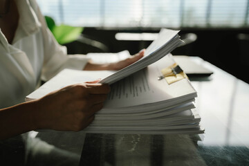 Close-up of businesswoman working at her desk with laptop and stack of documents, focusing on paperwork, reports and financial analysis in a busy office environment.
