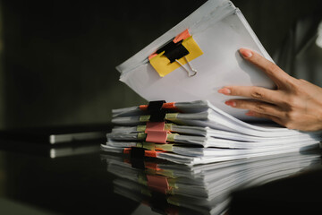 Close-up of businesswoman working at her desk with laptop and stack of documents, focusing on paperwork, reports and financial analysis in a busy office environment.