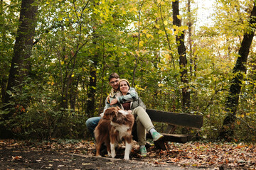 Couple sitting together on wooden bench with Australian Shepherd dog in colorful forest. Concept of love, peace, and connection