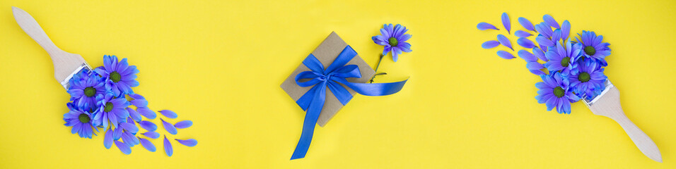 Gift box with tied blue bow and paintbrush with blue flowers on a yellow background. Top view.