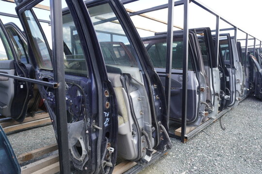 A row of car doors on metal racks. Outdoor car disassembly area, body parts, glass and metal, diagonal perspective, daylight.