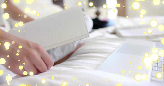 Holding female hand flipping lined notebook pages on bed, silver laptop nearby with yellow bokeh