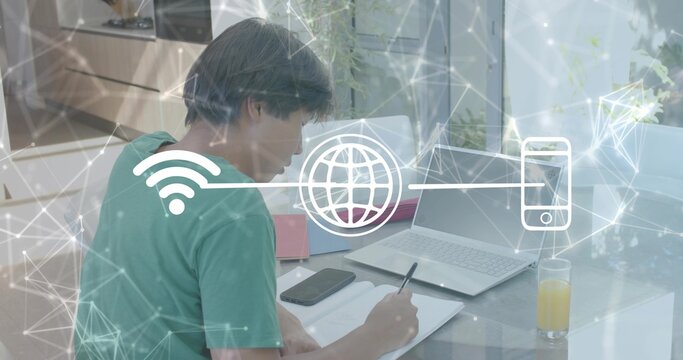 Asian man writing on notebook in home office, wearing green shirt, with laptop and network overlay