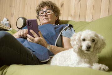 Senior woman sitting on her sofa at home with her white poodle using smartphone