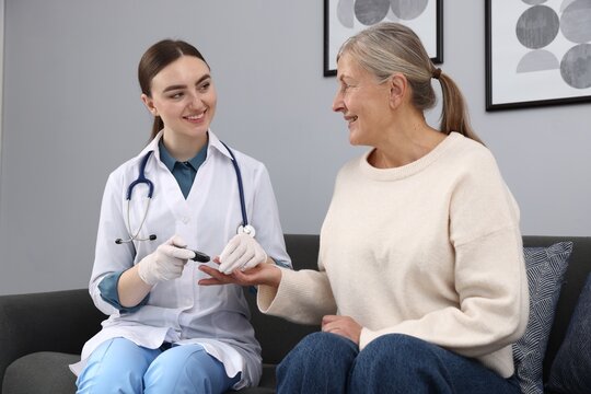 Diabetes. Smiling doctor in medical gloves checking patient's blood sugar level with glucometer on sofa at home