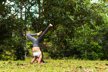 Athletic Latina woman performing an inverted yoga pose (headstand) outdoors. The scene conveys balance, strength, and a lifestyle focused on wellness and health.