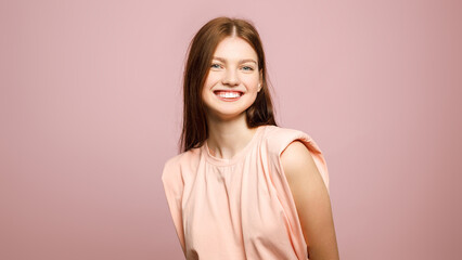 Obraz premium Cheerful young woman in a peach top smiling at camera against a pastel pink studio background with wide copy space.