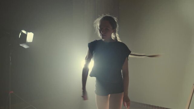 Skillful young woman in black clothes dancing modern contemporary choreography in a dark smoky studio with spotlights. A talented female performer expressing herself through dance and movement