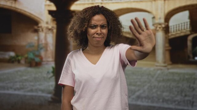 Woman holds hand up palm out in old town building courtyard, frowning and pulling face while pushing away with open palm; firm rejection.