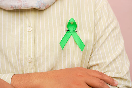 Woman wearing green ribbon on casual shirt against pink background, symbolizing mental health awareness, support, and solidarity.