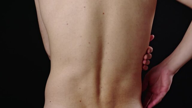 Close-up view of a shirtless person rubbing their painful lower back and hip, indicating discomfort, muscle strain, or injury against a solid black background, highlighting the spine and torso