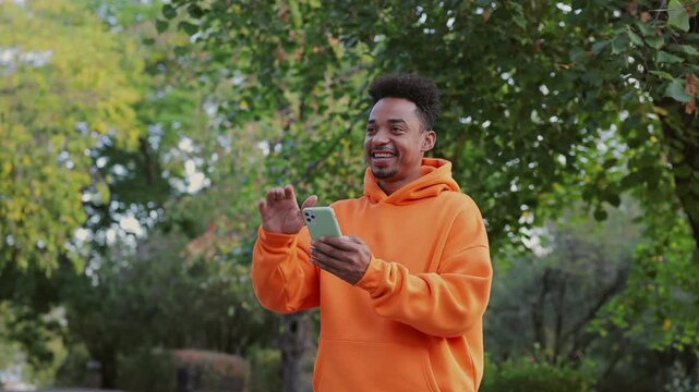 Young man wear orange hoody hold in hand use mobile cell phone waving hand greeting somebody walk rest relax in spring green city park go down sunshine alley outdoors on nature. Urban leisure concept