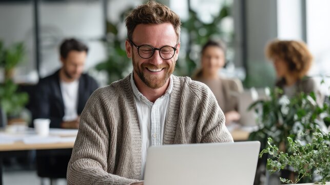 Happy bearded man with glasses using laptop in bright office setting. - Powered by Adobe