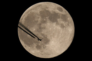 The moon and an airplane in the night sky. The plane is flying across the moon, against a black background.