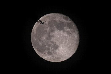 The moon and an airplane in the night sky. The plane is flying across the moon, against a black background.
