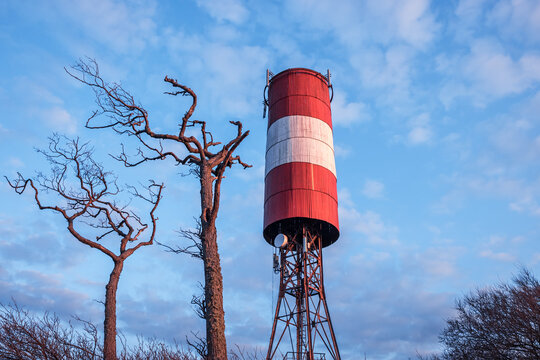 A tall red and white striped water tower stands among leafless trees against a bright blue sky. The scene evokes calm winter, rural industrial charm, and quiet everyday infrastructure