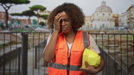 Woman technician in orange high visibility vest holding yellow hard hat with hand to forehead at ancient building ruins site; work stress.