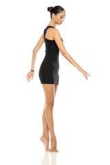 Studio shot of young tall woman with dark hair in bun wearing a black sleeveless dress, full length, performing a ballerina pose on white background.