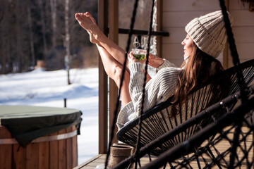 A woman in a chunky knit sweater and beanie relaxes on a porch hammock in a snowy setting, sipping a drink. The scene conveys warmth, comfort, and peaceful winter leisure