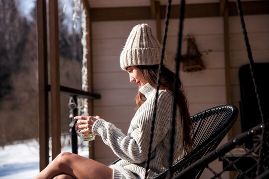 Woman Cozy Winter Relaxation On A Porch Hammock enjoy sipping hot tea with Knit Sweater And Beanie