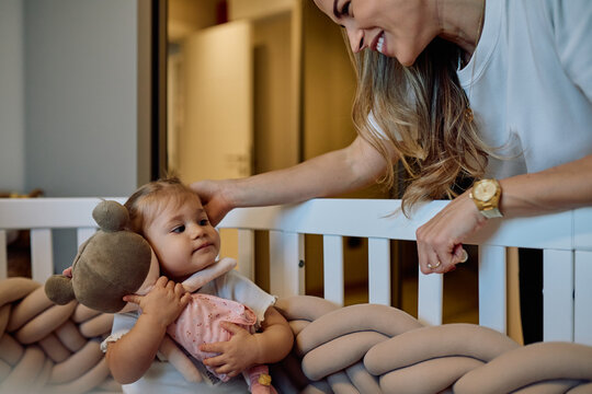 Baby holding doll in crib while mother gently strokes her head, tender moment of bonding, care and peaceful family connection