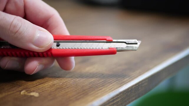 Hand Clicking Red Mini Stationery Knife on Wooden Desk Surface with Sharp Plastic Snap, Compact Utility Motion, Office Supply Control, Precision Blade Adjustment, Light Echo Sound, Practical Cutting