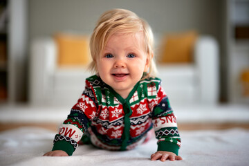 Cute Caucasian baby girl with bright blue eyes smiles widely while lying on her tummy in a cozy, festive Christmas holiday jumpsuit in a warm home setting.
