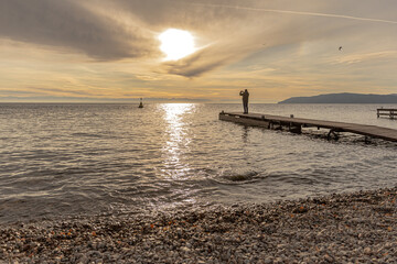 A Silhouette of lone figure stands on a wooden pier, capturing a sunset over a tranquil Baikal Lake. The pebble beach of the and distant hills. Taking photos of nature
