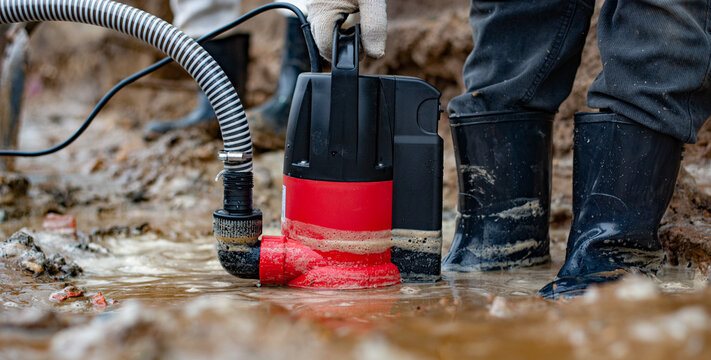 A drainage pump pumps dirty brown clay water out of a puddle. A man in the mud in rubber boots is standing nearby. High quality photo