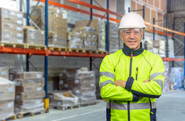 Warehouse worker with arms crossed wearing PPE and hard hat in industrial storage area.