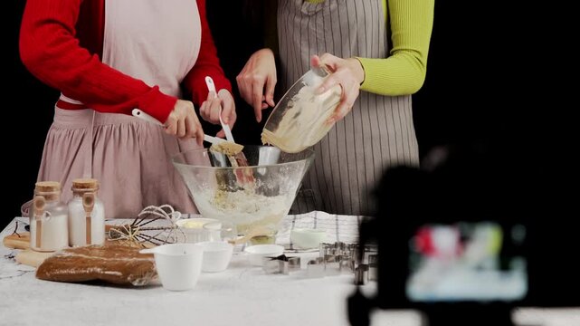 Two women scrape cookie dough into glass bowl of flour while baking together. Kitchen table with baking ingredients, utensils, and cookie cutters in frame