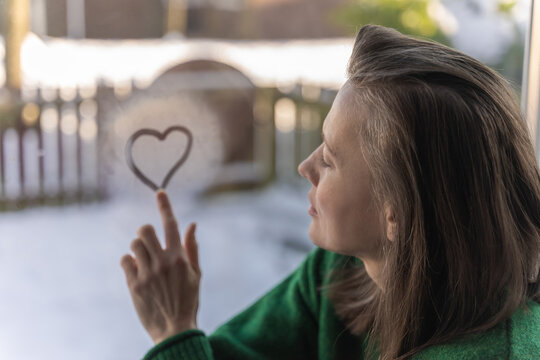 Woman drawing a heart with her finger on window - winter scene outside. Love and warmth concept, romantic winter mood. 