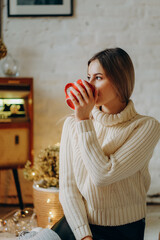 A young beautiful woman sits in a house near the window on Christmas Eve with a mug of hot tea.