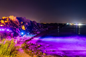 Magical night view of ancient fortress walls illuminated by blue and green lights on a beach, with the Mediterranean Sea and distant resort hotels. Side, Antalya, Turkey.

