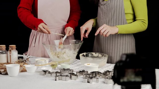 Two women in aprons mix cookie dough in glass bowl on kitchen table. Cookie cutters and baking ingredients are spread out, capturing cozy baking moment