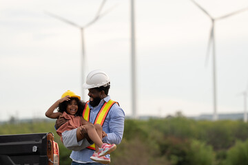 Engineer father carrying smiling daughter at wind turbine site, both wearing safety gear, representing love, support, STEM career inspiration, clean energy values, and strong family connection.
