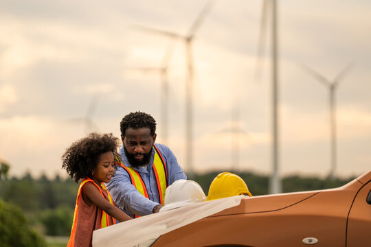 Engineer father and daughter wearing safety vests examining blueprint at wind turbine site, symbolizing STEM education, family bonding, renewable energy learning, and future career inspiration.