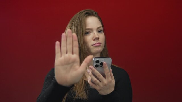 Woman raises palm and holds smartphone against red wall in studio with serious expression; rejection privacy.